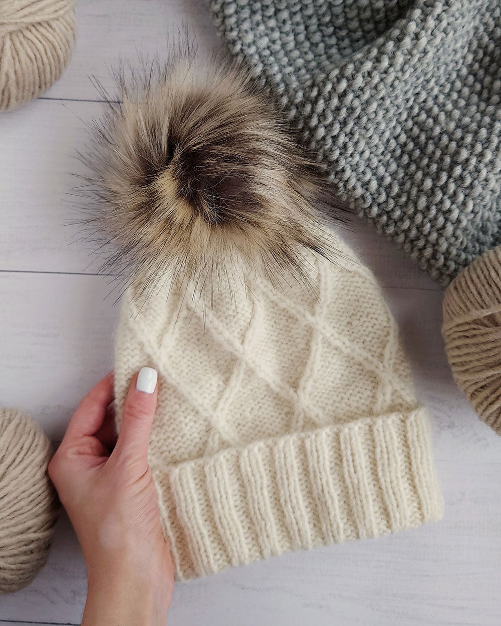 A cream-colored cable-knit beanie hat with a large brown and beige faux fur pom-pom, held by a hand with white nail polish against a light wood background with yarn balls and a gray knit blanket.
